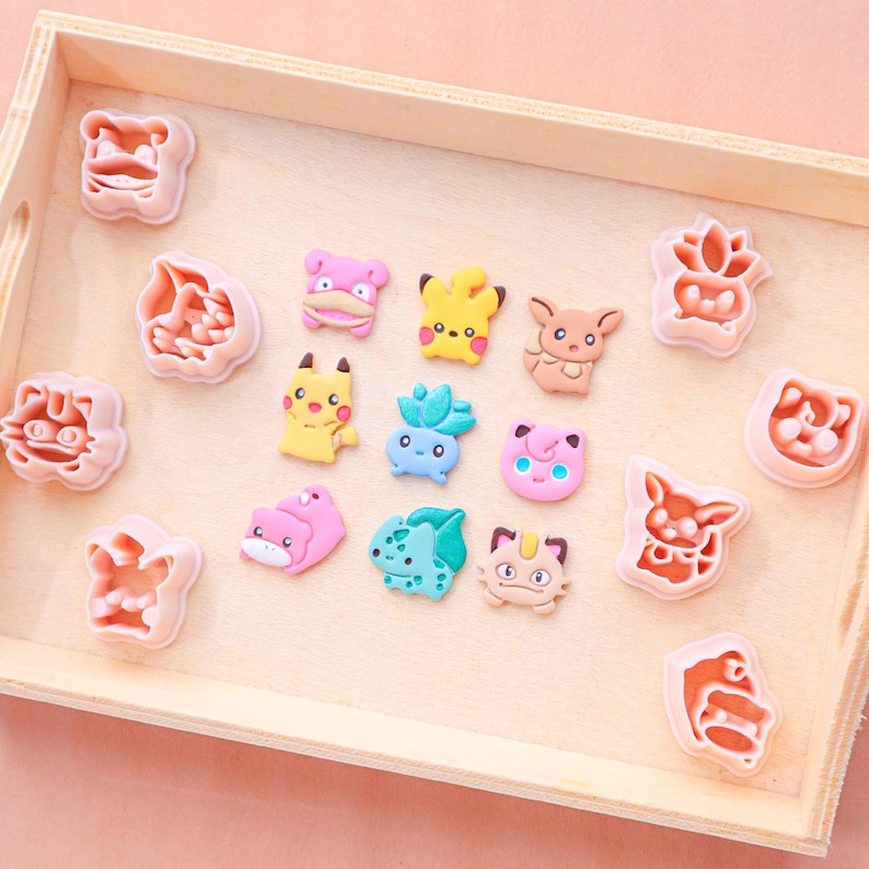 May include: A wooden tray holds an assortment of colorful character-shaped cookie cutters and matching character-shaped cookies. The cookie cutters are pink, and the cookies are yellow, pink, blue, and brown.