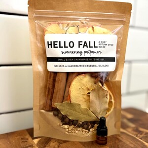 May include: A brown resealable pouch containing dried apple slices, cinnamon sticks, star anise, bay leaves, and spices. The label reads HELLO FALL A COZY AUTUMN SPICE BLEND simmering potpourri SMALL BATCH HANDMADE IN TENNESSEE INCLUDES A HANDCRAFTED ESSENTIAL OIL BLEND. A small amber glass dropper bottle is included.