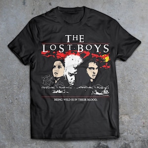 Peut inclure: T-shirt noir avec un graphique de trois personnages du film "The Lost Boys". Le texte "The Lost Boys" est imprimé en blanc au-dessus des personnages. Le texte "Being wild is in their blood." est imprimé en blanc en dessous des personnages.