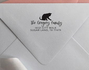 May include: A light blue envelope featuring a black ink stamp of a cat silhouette above the text The Gregory Family, 16121 CITY WALK, SUGAR LAND, TX 77479. The envelope is positioned with a pink envelope and a silver ribbon visible in the background.