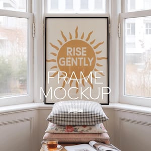 Op de afbeelding: Een ingelijste poster met de woorden "RISE GENTLY" en een oranje zonnegrafiek. De lijst is zwart. Onder het frame liggen gestapelde kussens met bloemen- en ruitpatronen. Een kopje thee en een tijdschrift liggen op een houten oppervlak.