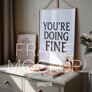 Può includere: Un mockup con due stampe artistiche incorniciate e oggetti decorativi su una cassettiera bianca. La cornice più grande mostra il testo "YOU'RE DOING FINE" in nero. Una cornice più piccola mostra un disegno a tratto di un fiore. Altri oggetti includono un flacone di profumo, un piattino e un taccuino.