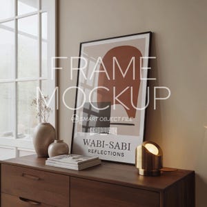 May include: A framed art print with the words "Wabi-Sabi Reflections" and abstract shapes in brown and beige tones. The print is displayed on a dark wood dresser, alongside a small lamp and vases. The scene evokes a minimalist aesthetic.
