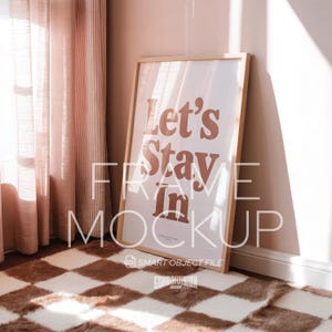 May include: A framed poster mockup with the text "Let's Stay In" in a warm brown font. The poster is leaning against a wall in a room with a patterned rug and pink curtains.