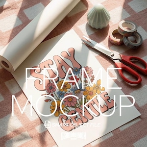 May include: A mock-up print with the words "STAY FRAME GENTLE" in a retro font, surrounded by colorful flowers. A roll of paper, scissors, decorative tape rolls, and a small shell-shaped object are also present.