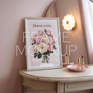 May include: A silver-framed print featuring a floral design and the words "bloom today". The print is propped on a white dressing table. Two lipsticks and a pink dish are also on the table, near a mirror.