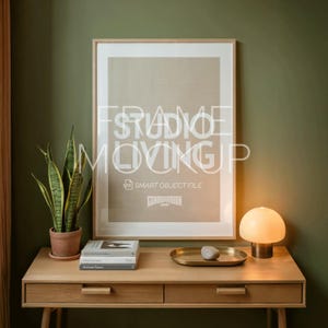 May include: A framed print with the text "STUDIO MOVING UP" and "SMART OBJECT FILE" is displayed above a wooden desk. A small lamp, books, a tray, and a potted snake plant are on the desk. The wall is painted a muted green.