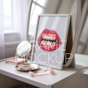 Könnte beinhalten: Ein weiß gerahmter Druck mit den Worten "Gloss Goals" über einem roten Lippenbild. Eine Gesichtswalze aus Rosenquarz, ein kleiner Spiegel und eine Schale mit Perlen liegen auf einer weißen Oberfläche. Das Bild ist ein Rahmen-Mockup.