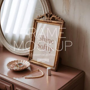 Puede incluir: Un marco de fotos dorado y ornamentado con el texto "Shine Softly" y "Frame Mock Up" sobre una superficie rosa. Un espejo redondo, un plato pequeño con pulseras, un collar de perlas y un pequeño objeto blanco también están en la superficie.