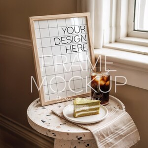 Puede incluir: Un marco de madera con el texto "YOUR DESIGN HERE" y "FRAME MOCKUP" está apoyado en una mesa pequeña y redonda. Una rebanada de pastel verde está en un plato blanco, junto a un vaso de café helado.