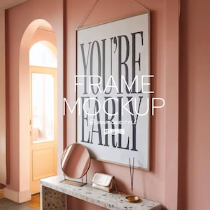 May include: A framed art print with the words "YOU'RE FRAME MOCKUP EARLY" in black lettering hangs on a pink wall. A small gold-framed mirror, a purse, and a decorative tray sit on a speckled table.