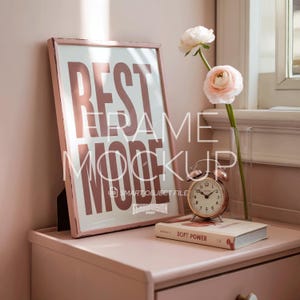 May include: A pink framed print with the words "REST MORE" in large letters. A small, vintage-style alarm clock and a book titled "SOFT POWER" sit on a pink nightstand. A vase with flowers is also present.