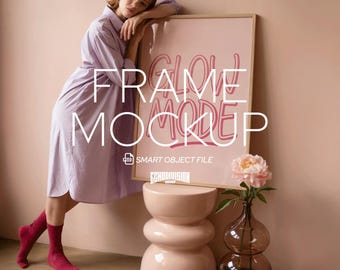 May include: A framed poster with the words "FRAME MOCKUP" in a handwritten style, leaning against a wall. A woman in a lavender dress leans against the frame. A pink stool and a glass vase with a flower are in front of the frame.