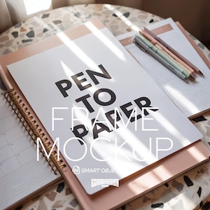 Puede incluir: Maqueta de papelería con un papel blanco con el texto negro "PEN TO FRAME PAPER MOCKUP". Descansa sobre un cuaderno rosa, junto a un cuaderno de espiral y varios bolígrafos. Los elementos están dispuestos sobre una superficie moteada.