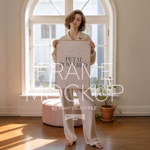 May include: A framed poster with the text "PETAL POWERED" and a line drawing. The frame is held by a person wearing a beige collared shirt and matching pants. The background includes a window, a pink ottoman, and a potted plant.