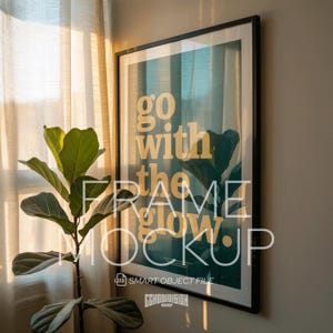 May include: A framed art print with the words "go with the flow" in a bold, yellow font, set against a teal and green background with a plant illustration. The frame is black. A potted plant is in the foreground.
