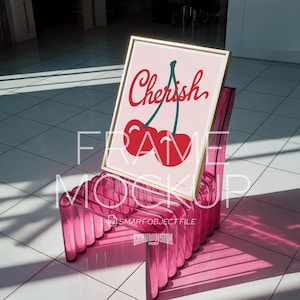 May include: A framed art print with a gold frame, featuring the word "Cherish" in red script above a pair of red cherries with green stems. The print is displayed on a transparent pink chair.