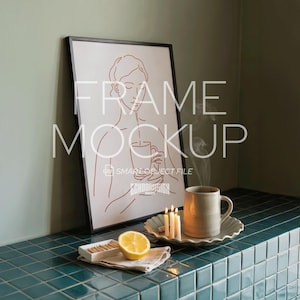 Könnte beinhalten: Ein gerahmter Kunstdruck mit einer minimalistischen Strichzeichnung einer Person, die eine Tasse hält. Eine Keramiktasse, Kerzen und eine Zitronenhälfte stehen auf einer türkisfarbenen Fliesenoberfläche. Die Worte "FRAME MOCKUP" sind aufgedruckt.