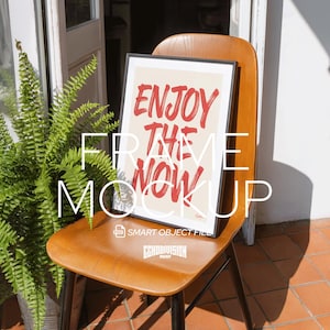 May include: A framed print with the words "ENJOY THE NOW" in red brush lettering. The framed art is displayed on a wooden chair. The image is a mockup, with the words "FRAME MOCKUP" in white text.