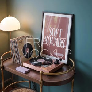 May include: A rose gold record player with a black vinyl record sits on a wooden bar cart. A stack of vinyl records and a framed poster with the words "SOFT SOUNDS" are also on the cart. A lamp with a white globe shade is in the background.