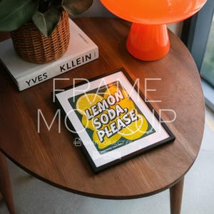 May include: A framed print with the text "LEMON SODA, PLEASE" in a retro style, featuring lemon slices on a yellow background. The print is in a black frame and rests on a wooden table. A book and an orange lamp are also visible.