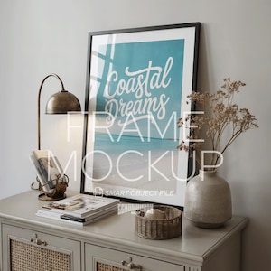 May include: A framed print with the words "Coastal Dreams" over a beach scene. The frame is black and rests on a gray cabinet with a lamp, magazines, a woven basket, and a vase with dried flowers. The lamp is brass-colored.