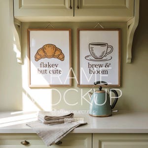 Könnte beinhalten: Zwei gerahmte Kunstdrucke mit Küchenthema. Ein Druck zeigt eine Croissant-Illustration mit dem Text "flakey but cute". Der andere Druck zeigt eine Kaffeetasse mit dem Text "brew & bloom". Ein türkisfarbener Wasserkocher und gefaltete Leinen-Servietten liegen auf der Theke.