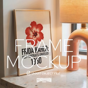 May include: A framed art print featuring a red hibiscus flower and text that reads "FRIDA KAHLO: SELF-PORTRAITS UNVEILED 1960". The gold frame rests on a speckled countertop, next to a peach-colored lamp.