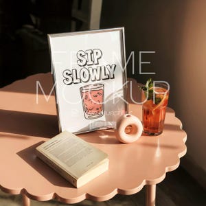 May include: A framed print with the words "SIP SLOWLY" and a cartoon glass of a pink drink. A glass of iced tea with mint and a small pink donut-shaped vase sit on a pink table next to a book.