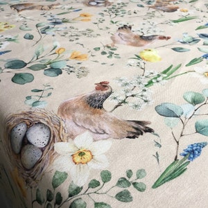 Könnte beinhalten: Eine beige Tischdecke mit einem floralen und Hühnerdruck. Die Tischdecke zeigt ein Nest mit Eiern, ein Huhn und verschiedene Blumen, darunter Narzissen, weiße Blüten und Blausternchen.