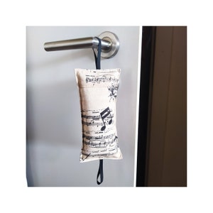 Può includere: Un fermaporta beige con un cinturino nero appeso a una maniglia della porta. Il fermaporta è realizzato in tessuto e presenta un design a nota musicale stampato.