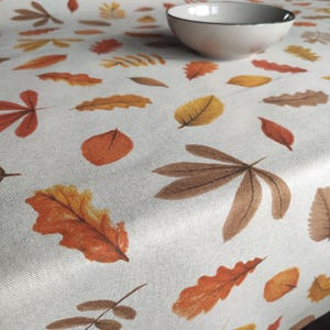 Op de afbeelding: Een crèmekleurig tafelkleed met een patroon van herfstbladeren in oranje, bruin en geel. Een witte kom met een zwarte rand staat op het tafelkleed. De bladeren zijn verschillend van grootte en vorm, wat een seizoensgebonden ontwerp creëert.