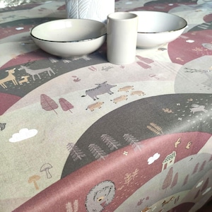 Könnte beinhalten: Eine Tischdecke mit einem sich wiederholenden Muster von Waldtieren, Bäumen und sanften Hügeln in Grau-, Rosa- und Beigetönen. Zwei weiße Schalen mit silbernen Rändern, eine weiße Vase und ein weißer Zylinder liegen auf der Tischdecke.