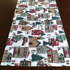 May include: White table runner with a repeating pattern of red, green, and brown houses, trees, and other festive details. The design is reminiscent of a winter village.