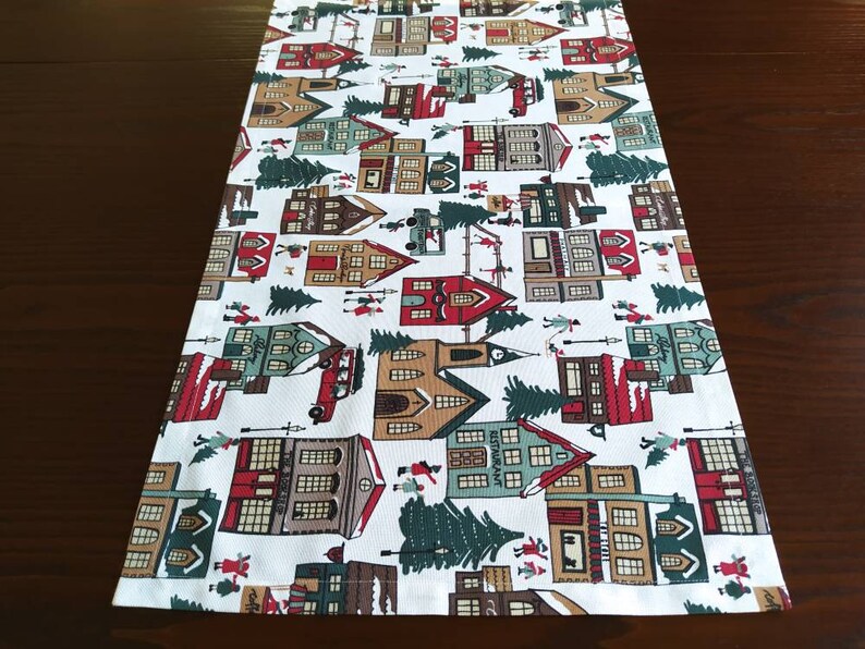 May include: White table runner with a repeating pattern of red, green, and brown houses, trees, and other festive details. The design is reminiscent of a winter village.