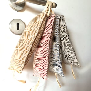 Könnte beinhalten: Fünf Türstopper aus Stoff in verschiedenen Farben, darunter Gold, Rot, Blau und Schwarz, jeweils mit einem geometrischen Rautenmuster. Sie hängen an einem Türgriff und zeigen ihr dekoratives Design.