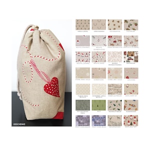 Op de afbeelding: Een beige stoffen cadeauzak met een rood hart en rode stiksels. De tas heeft het woord "Geschenke" gedrukt in de linkerbenedenhoek. De tas is omringd door verschillende stofstalen met verschillende kerstontwerpen.