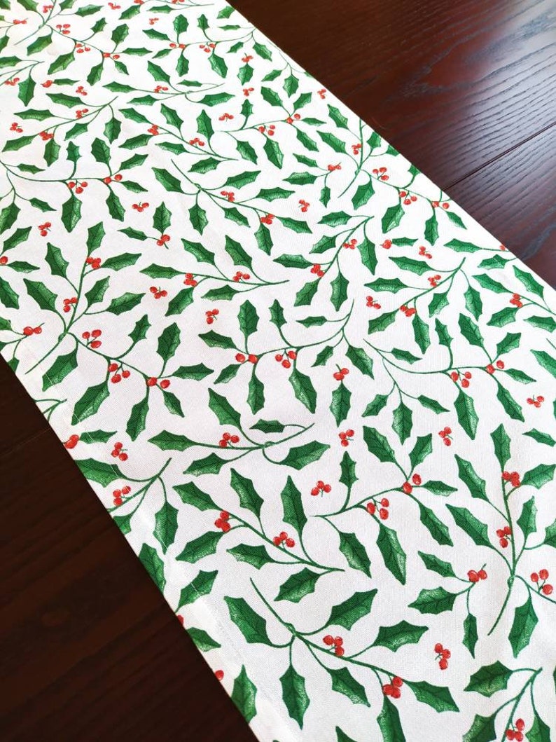 May include: White table runner with a repeating pattern of green holly leaves and red berries.