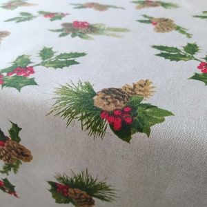 Peut inclure: Une nappe de couleur claire avec un motif répété d'illustrations sur le thème de Noël. Le motif comprend des pommes de pin, des feuilles de houx avec des baies rouges et des aiguilles de pin vertes. Le tissu a un aspect légèrement texturé.