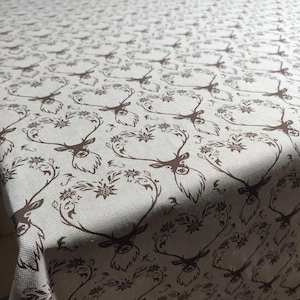 May include: A beige tablecloth featuring a repeating pattern of brown deer heads adorned with floral antlers. The fabric has a textured finish, and the design is consistently spaced across the surface.