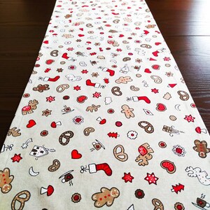 Könnte beinhalten: Ein beiger Tischläufer mit einem sich wiederholenden Muster aus Lebkuchenmännern, Brezeln, Sternen, Herzen und anderen weihnachtlichen Designs. Der Läufer ist aus einem weichen, gewebten Stoff gefertigt.