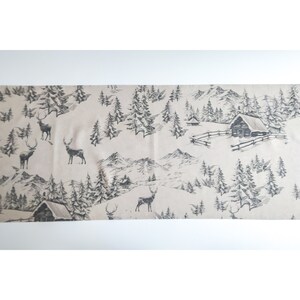 May include: A beige table runner with a black and white winter scene print. The print features trees, mountains, and deer.