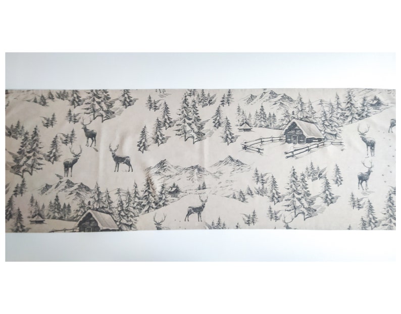 May include: A beige table runner with a black and white winter scene print. The print features trees, mountains, and deer.