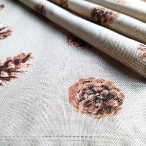 May include: A close-up of a beige fabric with a repeating pattern of brown pine cones. The fabric is folded in the upper right corner of the image.