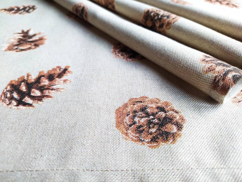 May include: A close-up of a beige fabric with a repeating pattern of brown pine cones. The fabric is folded in the upper right corner of the image.