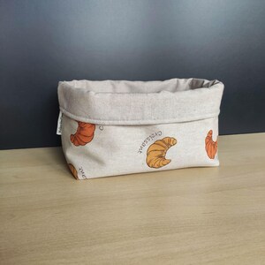 Può includere: Un cestino in tessuto beige con bordo ripiegato. Il cestino è decorato con illustrazioni di croissant arancioni e la parola "croissant". È appoggiato su una superficie di legno.