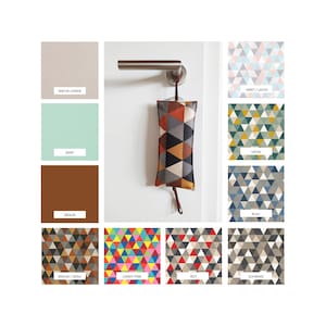 Puede incluir: Un tope de puerta con un patrón geométrico en tonos naranja, amarillo, negro y gris. El tope de puerta está colgado de una manija de puerta. El tope de puerta está disponible en 10 colores y patrones diferentes, incluyendo lino natural, menta, marrón, marrón/gris, rosa chicle, rojo, azul, verde, negro y menta/salmón.
