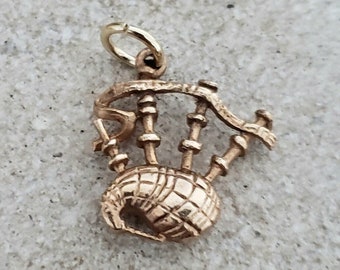 May include: A gold-colored charm in the shape of bagpipes. The charm has detailed features, including the chanter, drones, and bag. The charm is suspended from a small loop, suggesting it is designed to be attached to a necklace or bracelet. The charm is set against a neutral background.