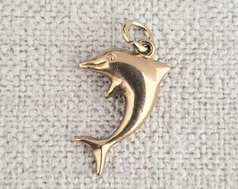 May include: A gold-colored dolphin charm pendant. The dolphin is leaping out of the water with its tail up.