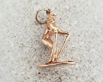 May include: A gold-toned charm of a skier in a downhill skiing pose. The charm is detailed with a skier wearing a hat and holding ski poles. The charm is on a white background.